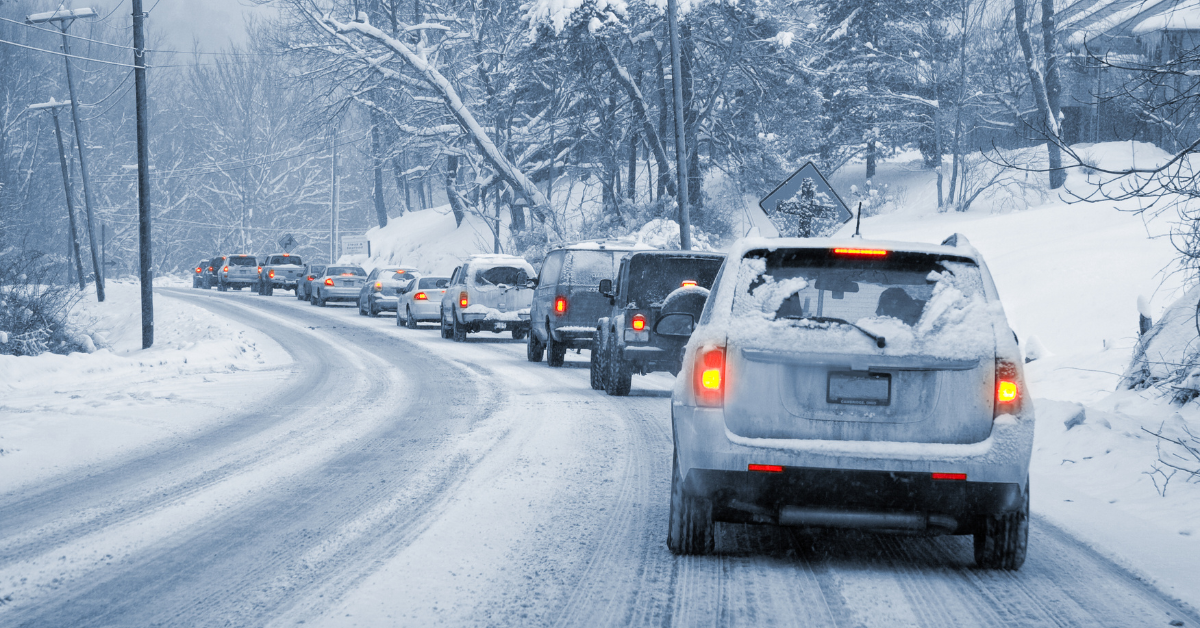 A line of cars drives slowly along a snow-covered road surrounded by trees and houses, with snow blanketing the ground, vehicles, and branches—perfect conditions to put Winter Cleaning Tips into practice for safer travel.
