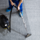 Person in blue pants and protective shoe covers uses a Carpet Cleaning Machine to clean a gray carpet; the difference between cleaned and uncleaned sections highlights the effectiveness of Carpet Cleaning.