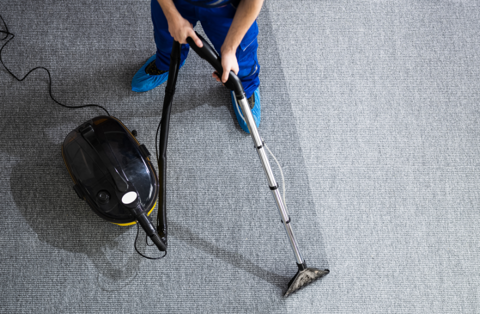 Person in blue pants and protective shoe covers uses a Carpet Cleaning Machine to clean a gray carpet; the difference between cleaned and uncleaned sections highlights the effectiveness of Carpet Cleaning.