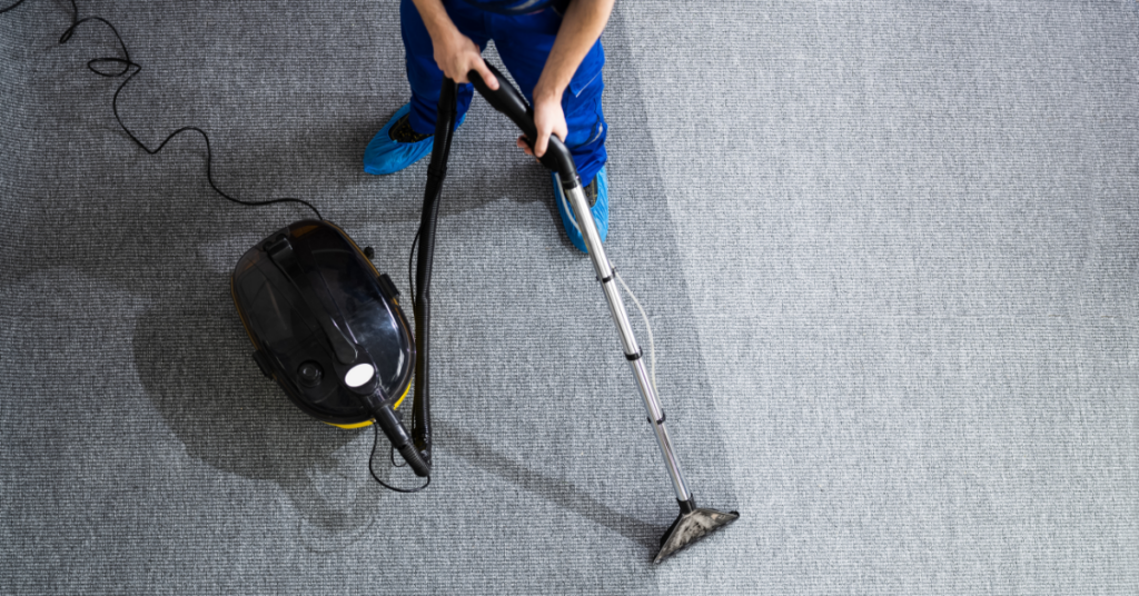 A person in blue clothing uses an at-home carpet cleaning machine to clean a gray carpet, creating a visible clean path. The scene is viewed from above.