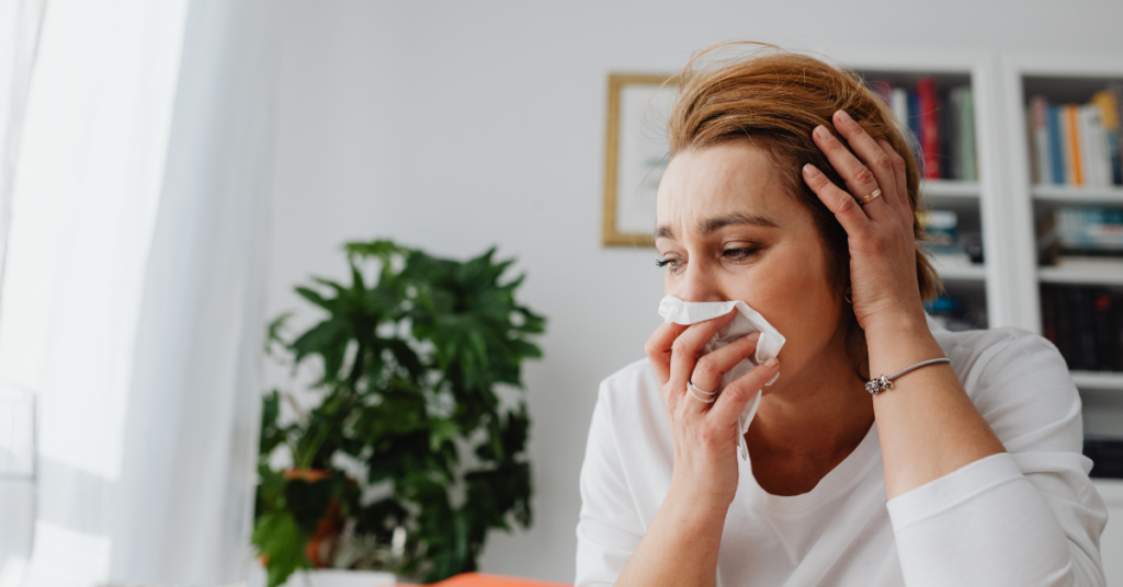 A woman sits indoors in Southern Indiana, holding a tissue to her nose and looking upset or unwell—likely affected by allergy season. One hand supports her head, with bookshelves and a green plant in the background, and wondering does steam cleaning carpet help with allergies.
