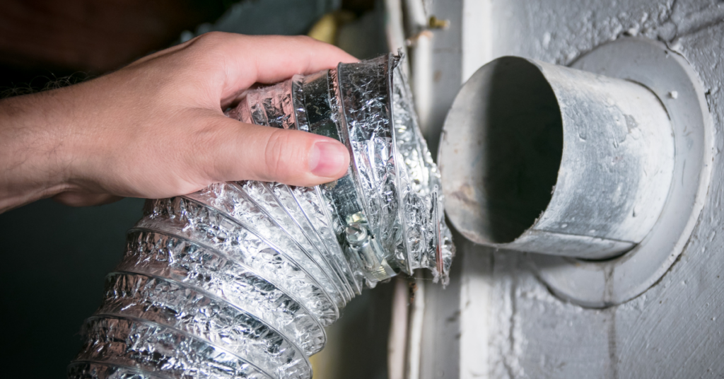 A hand is connecting a flexible silver dryer vent hose to a round metal wall duct, to complete an important step in dryer maintenance, an air duct and dryer vent cleaning, to help prevent fires and your dryer from taking too long.