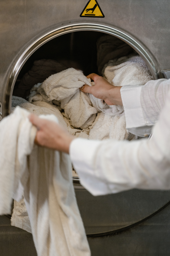Untitled design (4) A person wearing a white shirt is loading or unloading white towels from a large industrial washing machine with a warning sign above the door, showcasing standards similar to those seen in the best carpet cleaning facilities.