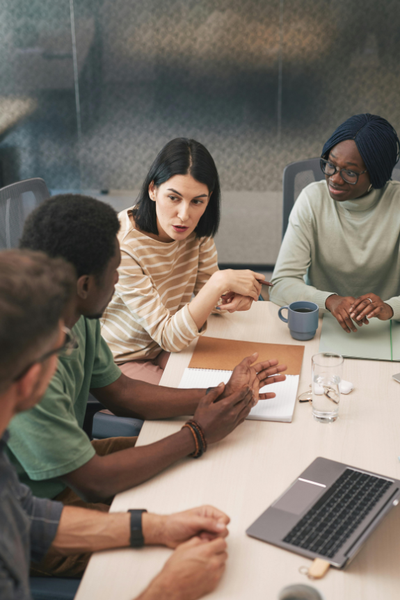 Untitled design (5) Four people sit around a table in a modern office, engaged in discussion about the best carpet cleaning methods. One woman gestures while speaking; others listen. Notebooks, a laptop, coffee mugs, and glasses are on the table.