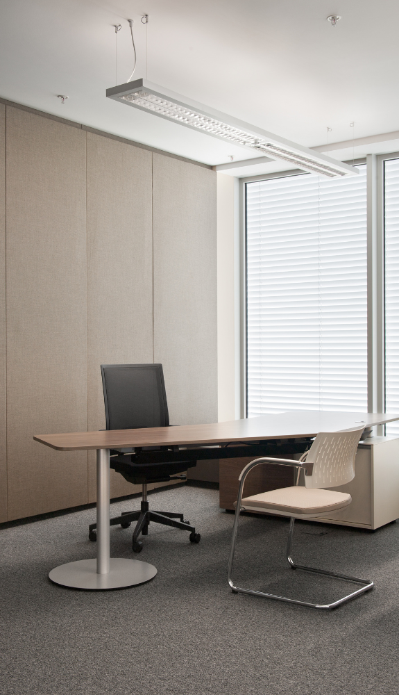 Untitled design (6) A modern office with a large wooden desk, black swivel chair, white visitor chair, best carpet cleaning for the carpeted floor, beige wall panels, and a window with white blinds allowing natural light in.