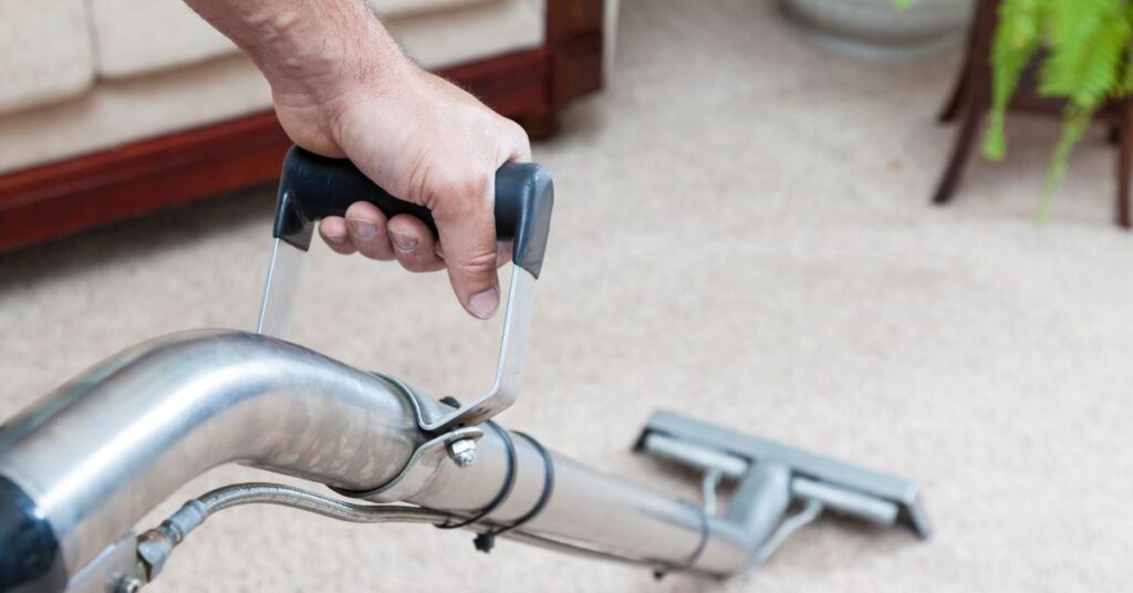 A close-up of a person’s hand holding the handle of a best carpet cleaning machine, moving it across a beige carpet in a living room with a sofa and plant in the background.