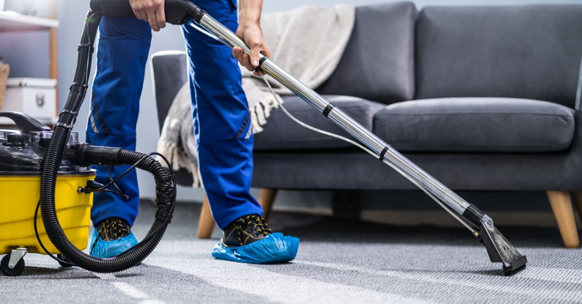 A person in blue pants and shoe covers uses a yellow carpet cleaning machine for the best carpet cleaning on a gray carpet in a living room with a gray sofa and a beige throw blanket in the background.
