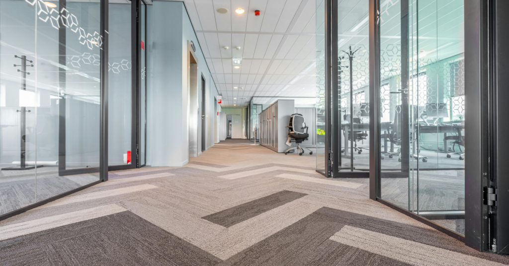 Modern office hallway with glass-walled meeting rooms and multicolored carpet, showing what office carpets look like with the proper commercial carpet maintenance.
