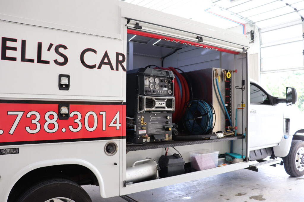 A white service van with an open side compartment reveals equipment, hoses, a generator, and various tools neatly organized inside—everything needed for professional carpet cleaning FAQ services. The red and white exterior displays partial company contact information.