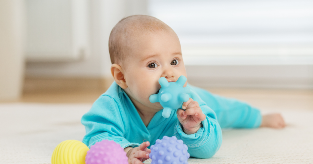 A baby in a blue onesie lies on the carpet floor, holding and chewing on a blue textured toy, highlighting the importance of safe carpet cleaning for families with young children.
