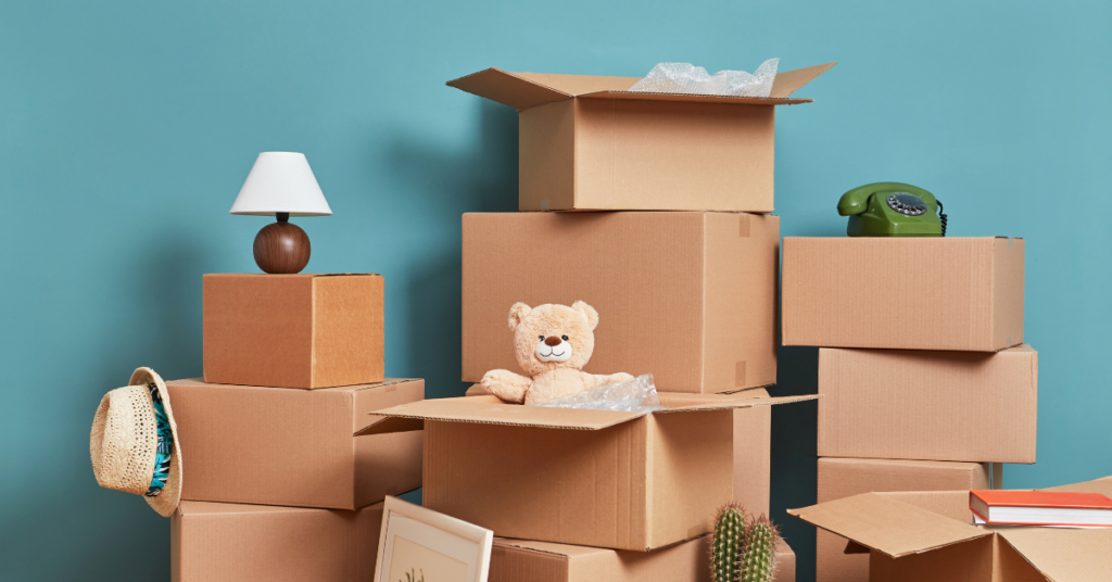 Several cardboard boxes are stacked against a blue wall. Some are open with a teddy bear and packing material inside, ready for move-out carpet cleaning or preparation for new tenants. Other items include a lamp, a green rotary phone, a hat, a framed picture, and a cactus—details property managers appreciate when ensuring seamless move-in carpet cleaning between residents.