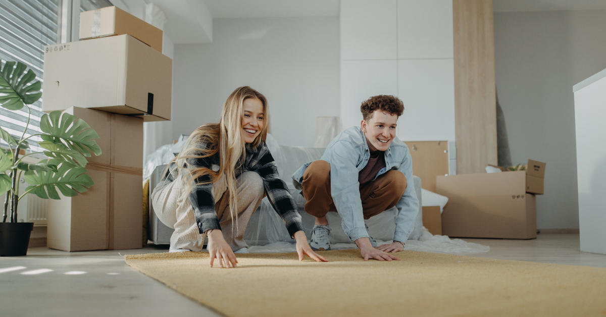 Two people smile as they lay out a yellow rug in a bright room filled with moving boxes, a large green plant, and sunlight streaming through the window—perfectly capturing that fresh feeling after move-in carpet cleaning.